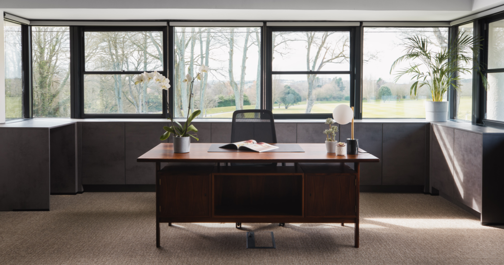 A desk in a Winslade Park office sits in front of windows, with lots of plants and desk is in command position facing door for Feng Shui